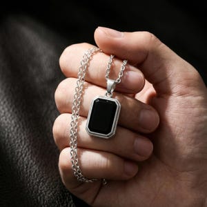 May include: A silver-toned pendant necklace featuring a rectangular black stone. The pendant has a double-layered frame and hangs from a delicate chain. The necklace is displayed against a dark background.