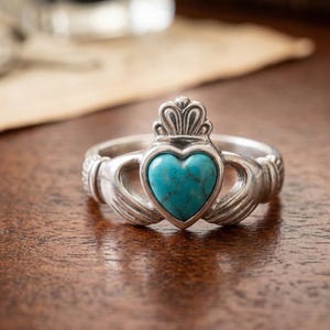 Puede incluir: Un anillo Claddagh de plata con una piedra turquesa en forma de corazón. El anillo presenta manos que sujetan el corazón, una corona encima y detalles intrincados. El anillo está sobre una superficie de madera.