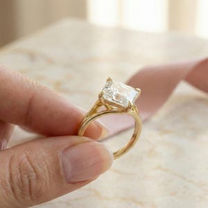 May include: A gold ring with a large, square-cut, clear gemstone. The ring is held by a hand, with a pink ribbon in the background. The ring's band is a simple, elegant design.