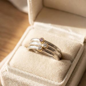 Puede incluir: Un conjunto de tres anillos apilables de plata, cada uno con una pequeña piedra preciosa. Las piedras preciosas incluyen una amatista púrpura y dos diamantes transparentes engastados en biseles de oro. Los anillos se muestran en un joyero de terciopelo beige.