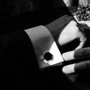 May include: Close-up of a black and white image of a cufflink on a white shirt cuff. The cufflink is a black, hexagonal shape with a silver border. The image is in black and white, with shadows and highlights.