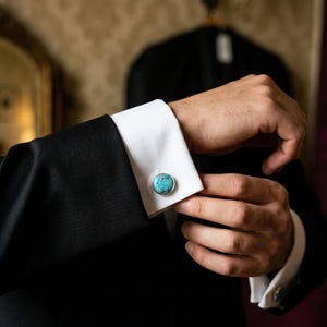 Op de afbeelding: Close-up van een persoon die een manchetknop op een zwart jasje aanpast. De manchetknop heeft een ronde, turquoise steen in zilver gezet. De manchet van het overhemd is wit.