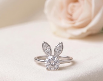 May include: A silver-toned ring featuring a central round-cut clear gemstone flanked by two marquise-cut clear gemstones arranged to resemble rabbit ears. The band is smooth and polished, showcasing a clean, metallic finish throughout the design.