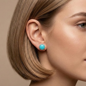 Peut inclure: Gros plan sur une oreille portant une boucle d'oreille ronde turquoise. La boucle d'oreille a un bord perlé argenté. La personne a les cheveux courts et bruns clairs et la peau claire. Le fond est de couleur neutre.