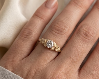 May include: A gold ring featuring a central round brilliant-cut clear gemstone. The band is crafted with detailed relief carvings of small roses and leaves on both sides of the center stone, creating a textured botanical design around the finger.