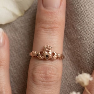 Op de afbeelding: Een roségouden Claddagh-ring, een traditioneel Iers ontwerp. Het heeft een hart, handen en een kroon, symbolen van liefde, loyaliteit en vriendschap. De ring wordt op een vinger getoond.