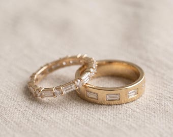 May include: Two gold rings are displayed together. One is a thin band featuring a continuous row of baguette-cut diamonds. The second is a wider, smooth gold band inlaid with a series of rectangular baguette-cut diamonds spaced evenly around the ring.