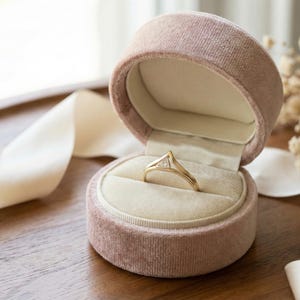 May include: A gold ring with a triangular diamond sits in an open pink velvet ring box. The ring box has a cream-coloured interior. The box is on a wooden surface, with a white ribbon nearby. The ring is a symbol of commitment.