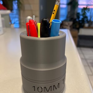 Puede incluir: Un organizador de escritorio gris con forma de llave de vaso, con el texto "10MM" en el lateral. Contiene un lápiz amarillo, bolígrafos rojos y negros y un rotulador azul. El organizador está sobre una superficie blanca.