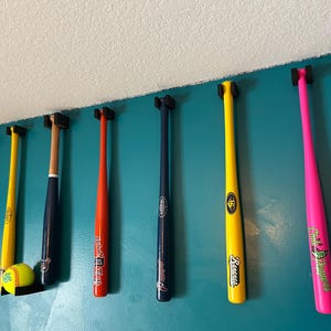 May include: A collection of baseball bats in various colors, including red, yellow, orange, navy, and pink, mounted on a teal wall. A yellow softball rests on a black shelf. The bats are displayed for decorative purposes.