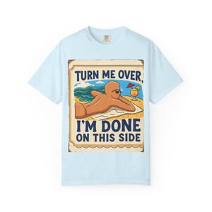 Op de afbeelding: Een lichtblauw t-shirt met een afbeelding van een peperkoekkleurig figuur dat op een strandlaken ligt. De tekst luidt "TURN ME OVER, I'M DONE ON THIS SIDE", vergezeld door een illustratie van een tropisch drankje met een parasolletje.