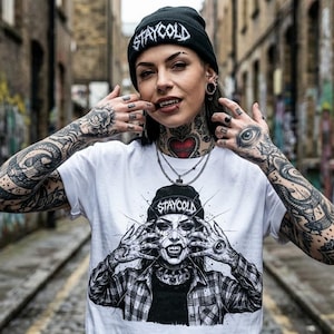 Puede incluir: Una camiseta blanca de manga corta con un estampado gráfico en blanco y negro de una persona con un gorro con el texto STAYCOLD, que se sujeta la cara con ambas manos y lleva una camisa de cuadros. El gráfico también incluye el texto STAYCOLD.