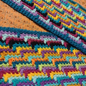 May include: Close-up of a colorful, hand-crocheted scarf with a geometric pattern. The scarf features a vibrant mix of colors including teal, purple, yellow, orange, and black. The edges are finished with a teal border, showcasing the detailed craftsmanship.
