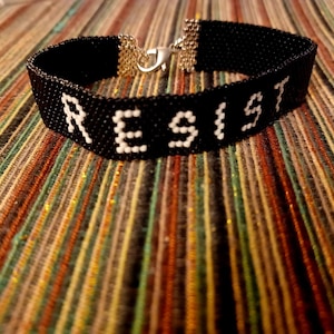 May include: Black beaded bracelet with the word "RESIST" spelled out in white beads. The bracelet has a silver clasp and is displayed on a colorful striped surface. A statement piece.