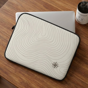 May include: Cream-colored laptop sleeve with a topographic map design and a compass rose emblem. The sleeve has a black border and a zipper closure. A laptop and a mug of coffee are also visible.
