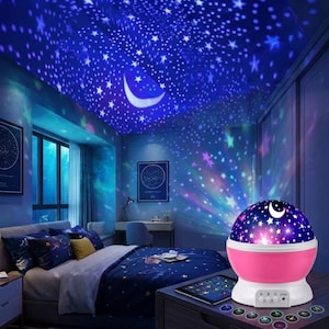 May include: A pink and white star projector lamp projects a starry night sky onto a bedroom ceiling. The lamp has a dome with stars and a crescent moon. The room has a bed with a blue comforter and pillows, and a window.