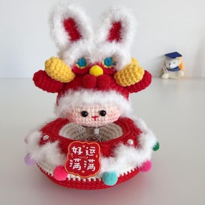 May include: A handmade, red and white crochet bunny toy with yellow accents and a smiling face. The toy has a spring base and a decorative red ring with the text "Good Luck" in Chinese. A small graduation bear is in the background.