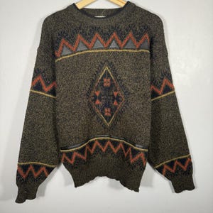 Könnte beinhalten: Ein olivgrüner Vintage-Pullover mit geometrischen Mustern. Der Pullover hat ein rautenförmiges Design in der Mitte und Zickzackmuster in Orange und Schwarz. Der Pullover hat einen Rundhalsausschnitt und lange Ärmel.
