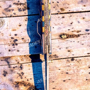 May include: A decorative folding knife with a patterned handle featuring blue, gold, and silver accents. The blade is extended, and the knife is resting on a weathered wooden surface. The text "AS BLADES" is visible.