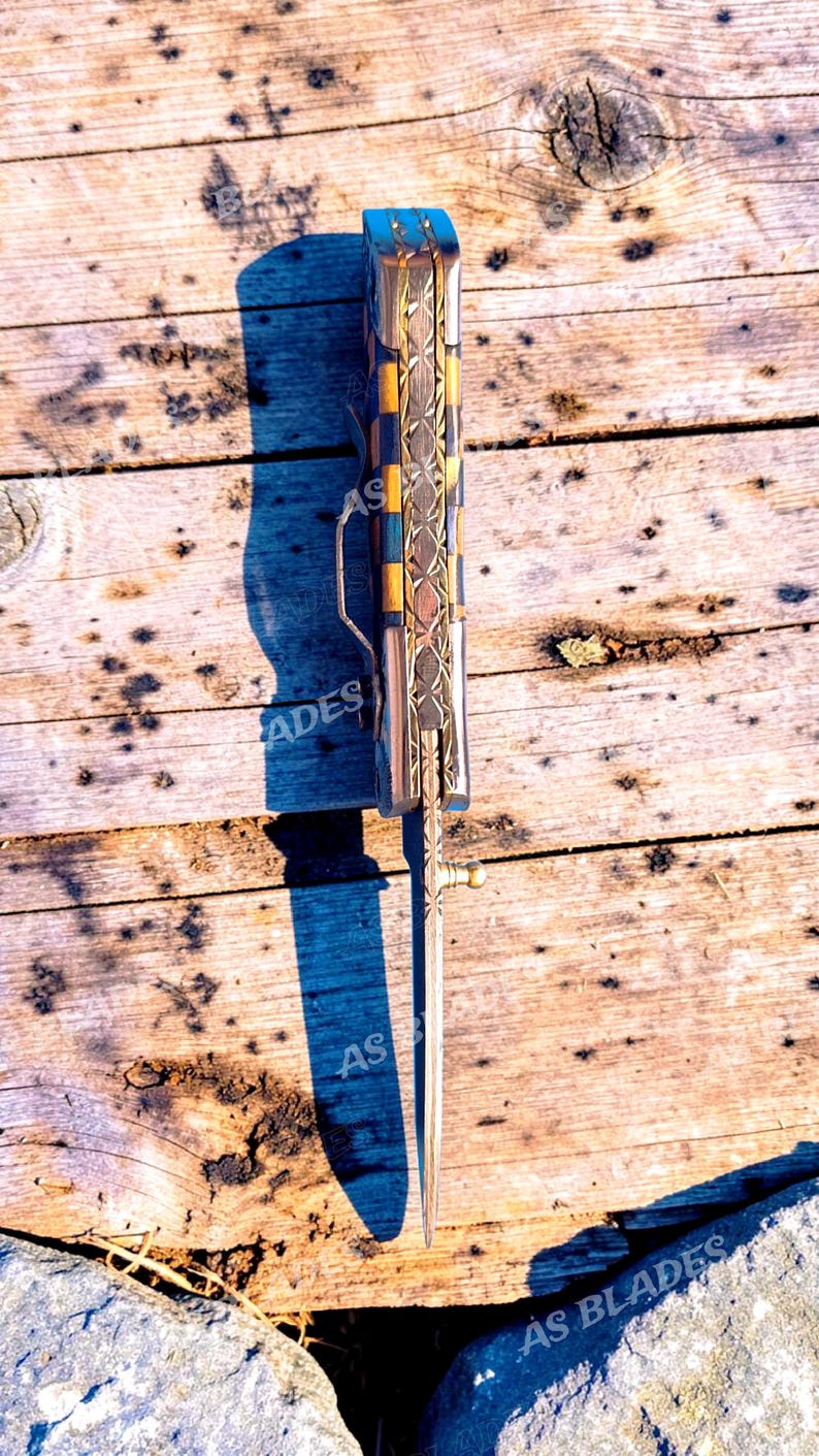 May include: A decorative folding knife with a patterned handle featuring blue, gold, and silver accents. The blade is extended, and the knife is resting on a weathered wooden surface. The text "AS BLADES" is visible.