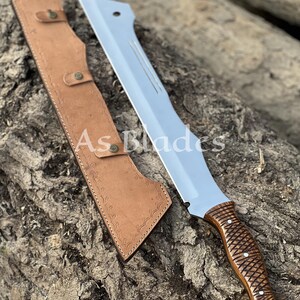 May include: A large, silver-bladed machete with a brown, textured handle and a matching leather sheath. The blade has a straight edge and a pointed tip. The sheath is tan and has a decorative pattern. The text "As Blades" is visible.