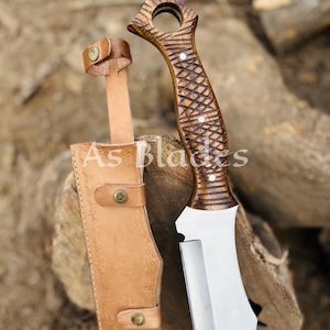 May include: A handcrafted knife with a wooden handle and a stainless steel blade, accompanied by a leather sheath. The handle features a detailed carved design. The sheath is tan with decorative stitching and brass-colored rivets. The text "As Blades" is visible on the sheath.