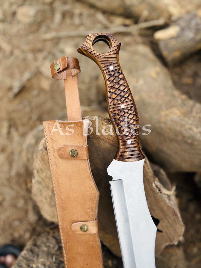 May include: A handcrafted knife with a wooden handle and a stainless steel blade, accompanied by a leather sheath. The handle features a detailed carved design. The sheath is tan with decorative stitching and brass-colored rivets. The text "As Blades" is visible on the sheath.