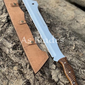 May include: A large, silver-bladed machete with a brown, textured handle and a matching leather sheath. The blade has a straight edge and a pointed tip. The sheath is tan and has a decorative pattern. The text "As Blades" is visible.