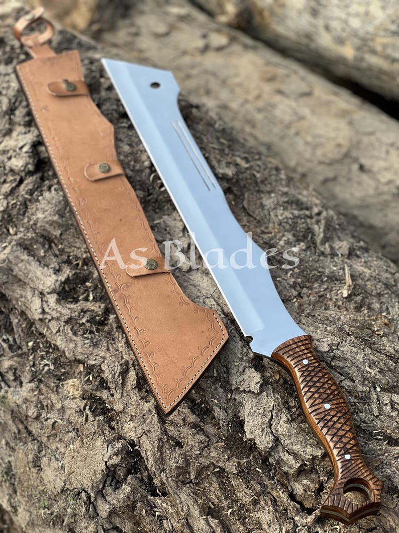 May include: A large, silver-bladed machete with a brown, textured handle and a matching leather sheath. The blade has a straight edge and a pointed tip. The sheath is tan and has a decorative pattern. The text "As Blades" is visible.