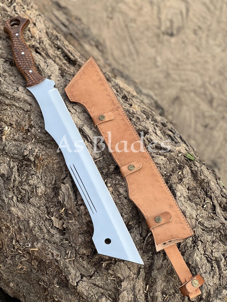 May include: A large, silver blade with a brown wooden handle and a leather sheath. The blade has a unique shape with a pointed tip and several grooves. The sheath is tan and has decorative stitching and button closures. The text "As Blades" is visible.