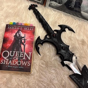 以下が含まれることがあります： サラ・J・マース著『Queen of Shadows』の赤い表紙の本と、装飾が施された柄、銀色の刃、赤い宝石のアクセントが付いた黒い鞘を持つ、黒い装飾用の剣が並んでいます。表紙には剣を持つキャラクターが描かれています。