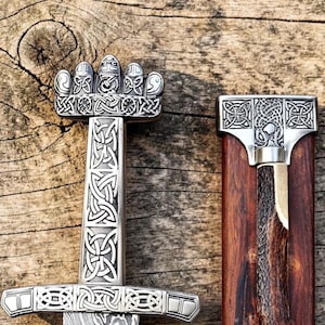 Pode incluir: Imagem detalhada de uma espada e bainha viking. A espada apresenta uma empunhadura prateada ornamentada com intrincados nós e uma lâmina estampada. A bainha é feita de madeira com detalhes prateados.