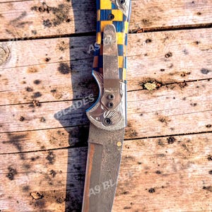 May include: A folding knife with a patterned handle in blue and yellow, and a silver blade. The knife has the text "AS BLADES" on the blade and handle. The knife is lying on a wooden surface.