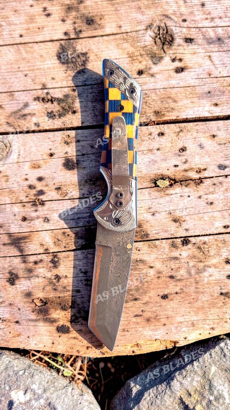 May include: A folding knife with a patterned handle in blue and yellow, and a silver blade. The knife has the text "AS BLADES" on the blade and handle. The knife is lying on a wooden surface.