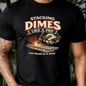 May include: Black t-shirt with a graphic featuring the text "STACKING DIMES LIKE A PRO" in orange and white. The design includes a welding torch and a metal plate with a weld, with the phrase "one bead at a time" below.