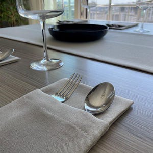 May include: A dining table setting with a light gray linen napkin holding a silver fork and spoon. A black bowl and wine glasses are visible on a light gray table runner. The table is set near a window.