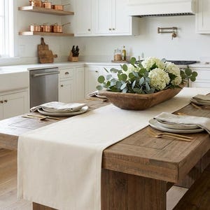 Könnte beinhalten: Ein rustikaler Esstisch aus Holz mit einem cremefarbenen Tischläufer, gedeckt mit Tellern, Servietten und goldfarbenem Besteck. Eine Holzschale mit weißen Blumen und Grünzeug steht in der Mitte. Die Küche hat weiße Schränke und Kupferkochgeschirr.