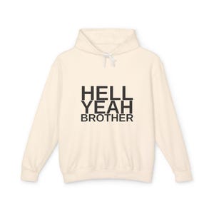 May include: A cream-coloured long-sleeve hooded sweatshirt featuring the black text HELL YEAH BROTHER printed in a bold, sans-serif font across the centre of the chest. The garment includes a matching hood with white drawstring cords.