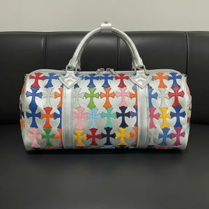 Könnte beinhalten: Silberne Reisetasche mit einem sich wiederholenden Muster aus bunten Kreuzen. Die Tasche hat einen silbernen Griff und Zierleisten. Die Kreuze sind in verschiedenen Farben, darunter Rot, Blau, Grün und Rosa. Die Tasche liegt auf einer schwarzen Oberfläche.