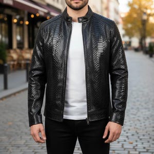 May include: A black leather jacket with a snakeskin texture, featuring a stand-up collar and a front zipper. The jacket is worn over a white t-shirt and paired with black pants. The image is taken outdoors.