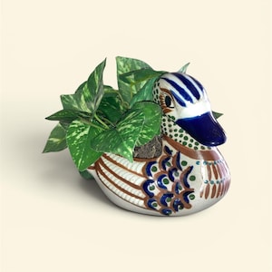 May include: A ceramic duck-shaped planter with a glossy white finish, featuring hand-painted blue, brown, and green decorative patterns. The duck has a solid blue beak and contains a green leafy plant with variegated foliage emerging from its back.