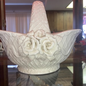 May include: Cream-colored ceramic basket with a handle, featuring a diamond-patterned design and three-dimensional rose embellishments. The basket has a wide, open shape and is suitable for decorative purposes.