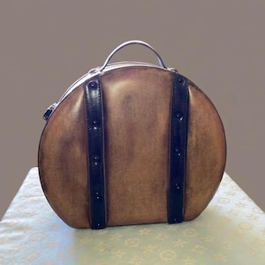 May include: A round, tan-colored bag with a distressed finish, featuring two vertical black leather-like straps adorned with dark studs. The bag includes a top handle and a zipper closure, presenting a structured, circular silhouette for carrying items.