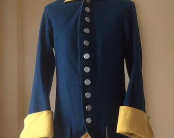 May include: A navy blue historical coat with a yellow collar and cuffs. The coat features a row of silver buttons down the front and yellow lining visible at the hem. The garment is displayed on a mannequin, showcasing its tailored design.