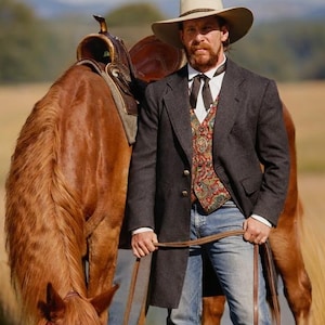 Puede incluir: Un hombre con sombrero de vaquero, chaleco y abrigo largo está junto a un caballo marrón en un campo. El caballo lleva silla y brida. El hombre sostiene las riendas. La escena evoca un tema del Oeste.