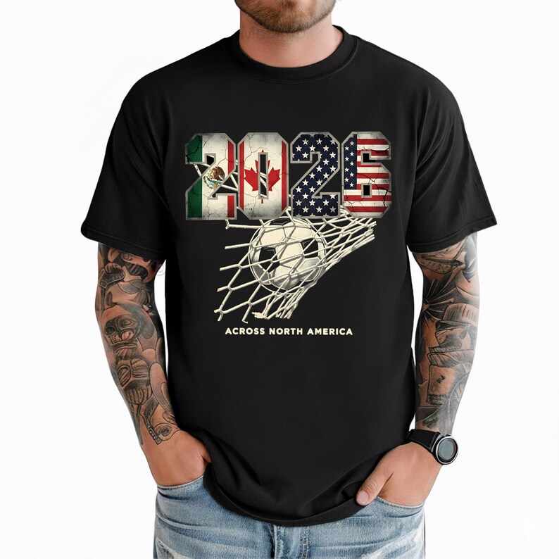 Op de afbeelding: Zwart t-shirt met het jaar "2026" in een ontwerp met de vlaggen van Mexico, Canada en de Verenigde Staten. Een voetbal zit in een net, met de tekst "ACROSS NORTH AMERICA" eronder.