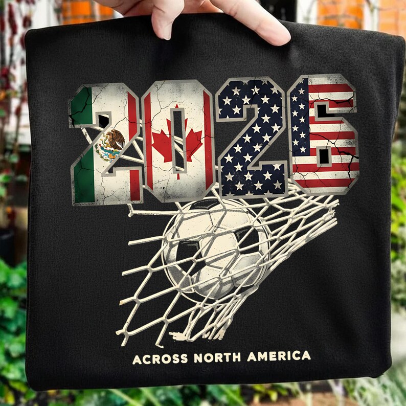 Op de afbeelding: Zwart t-shirt met het jaartal "2026" met de vlaggen van Mexico, Canada en de Verenigde Staten. Een voetbal zit in het net. De tekst "ACROSS NORTH AMERICA" staat onderaan.