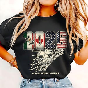 Op de afbeelding: Zwart t-shirt met het jaartal "2025" weergegeven met de vlaggen van Mexico, Canada en de Verenigde Staten. Een voetbal zit in het net, met de tekst "ACROSS NORTH AMERICA" eronder.