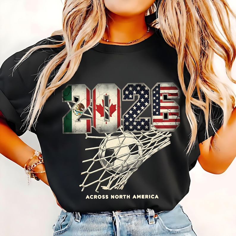 Op de afbeelding: Zwart t-shirt met het jaartal "2025" weergegeven met de vlaggen van Mexico, Canada en de Verenigde Staten. Een voetbal zit in het net, met de tekst "ACROSS NORTH AMERICA" eronder.