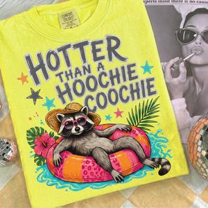 Op de afbeelding: Geel Comfort Colors T-shirt met de tekst "Hotter Than a Hoochie Coochie" in grijze letters. De afbeelding toont een wasbeer met zonnebril en strohoed, liggend op een opblaasbare ring. Tropische bloemen en sterren omringen de wasbeer.